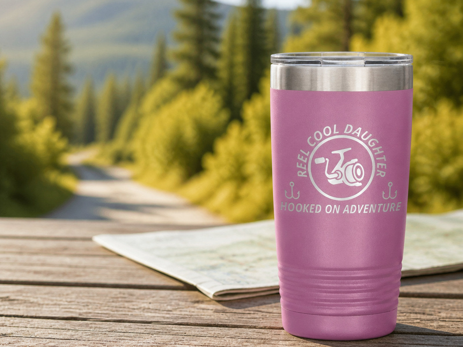 Light-purple 20-ounce REEL COOL DAUGHTER engraved tumbler on a picnic table overlooking a forest road.