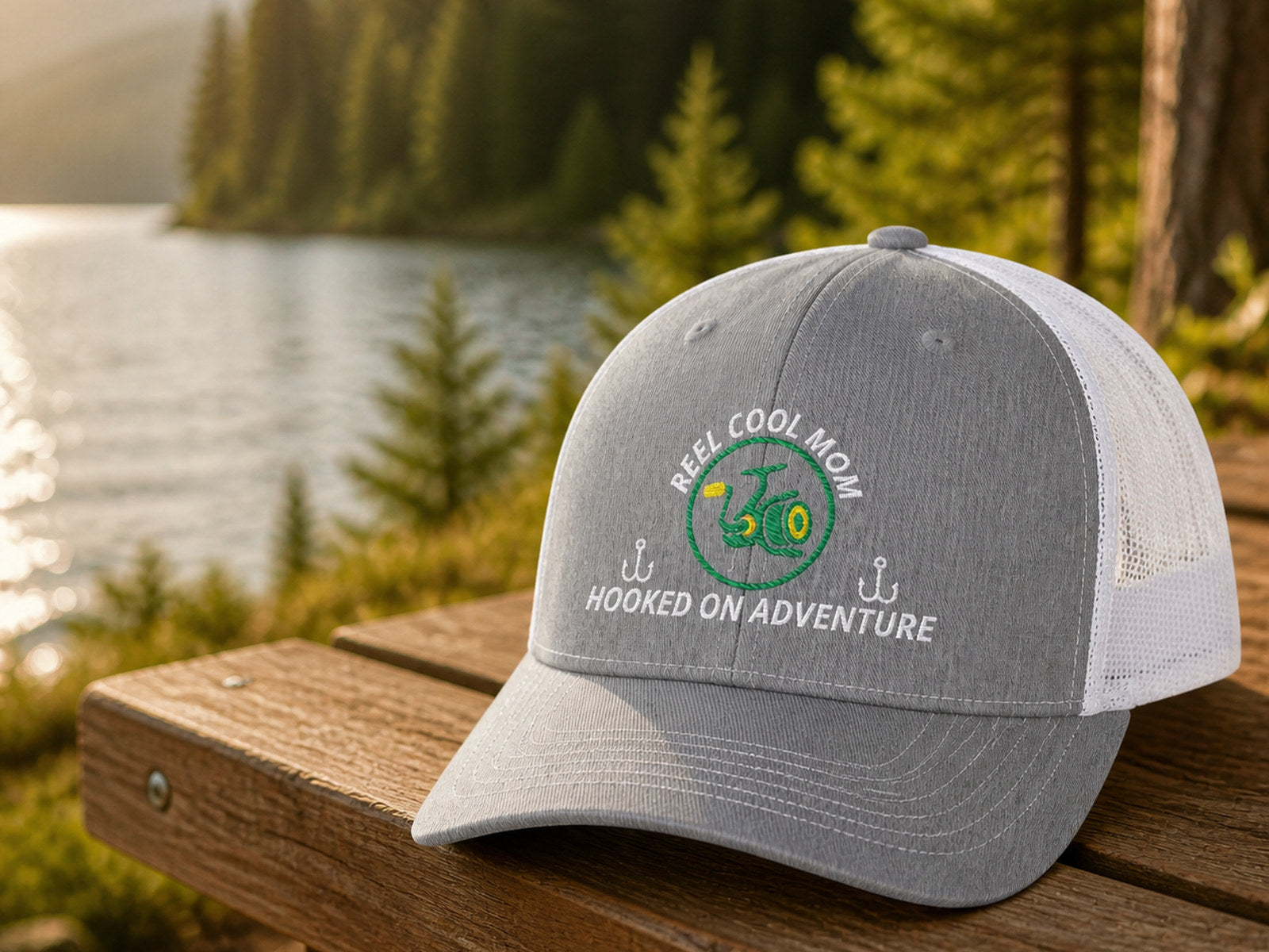 Heather-gray-and-white REEL COOL MOM embroidered trucker cap on a lakeside wooden bench.