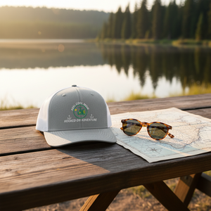 REEL COOL MOM trucker cap with heather gray front and white mesh back on a wooden table in a warm lakeside setting.
